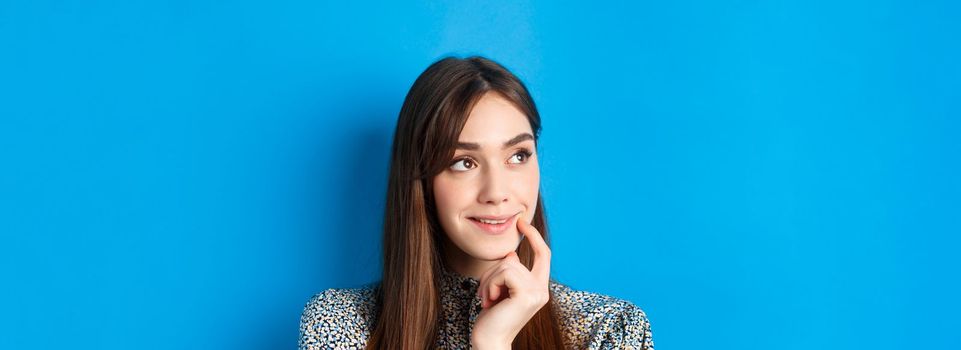 Interesting. Pensive young girl looking aside at logo and smiling, touching lip thoughtful, have an idea, standing on blue background.