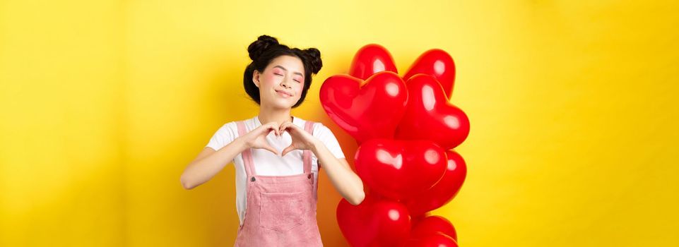 Valentines day concept. Cute asian girl dreaming of romance, close eyes and showing heart gesture, smiling happy, standing near red romantic balloons, yellow background.