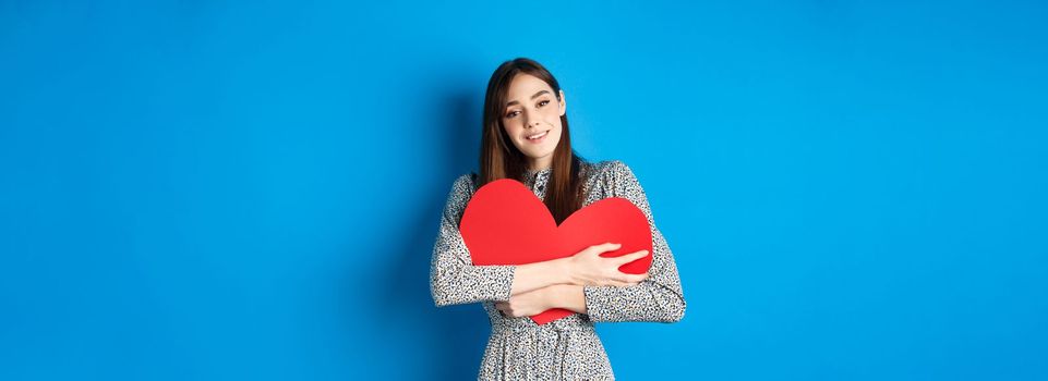 Valentines day. Dreamy romantic woman hugging big red heart cutout, looking sensual at camera, standing in dress on blue background.