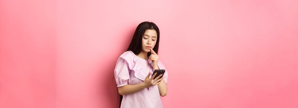 Online shopping. Pensive asian girl looking at smartphone screen, making choice, standing in dress against pink background.