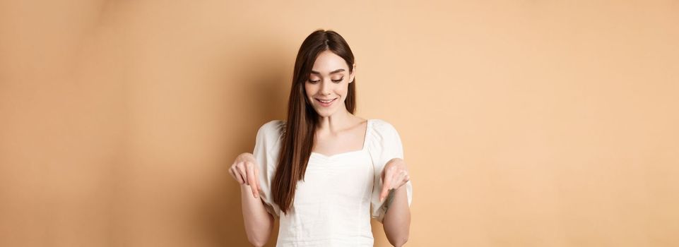 Smiling beautiful female model looking and pointing down with pleased happy face, showing something romantic, standing on beige background.