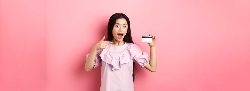 Happy cute asian woman showing plastic credit card, recommending bank special deal, standing against pink background.