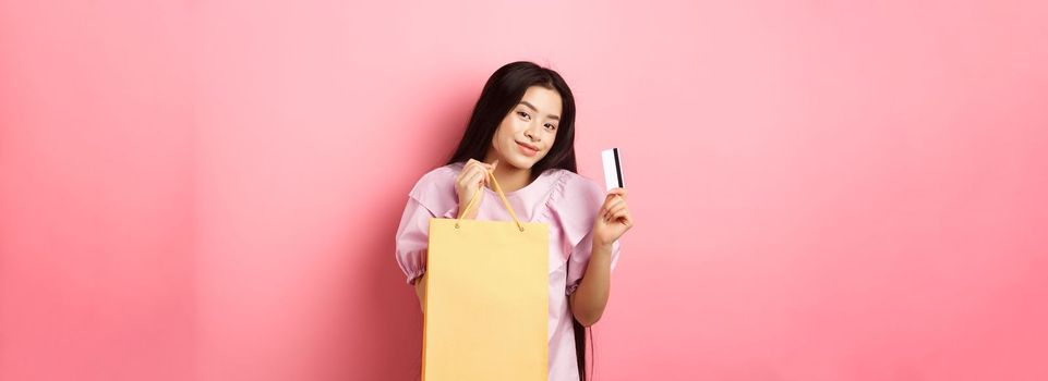 Shopping. Stylish asian girl showing shop bag and plastic credit card, standing on pink background.