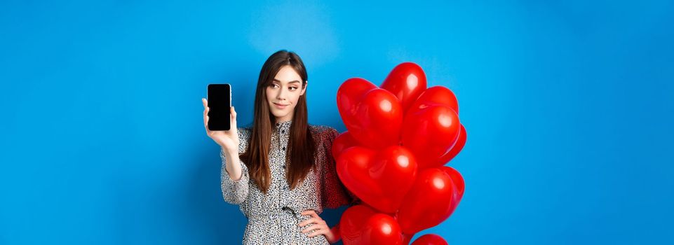 Valentines day. Pretty smiling woman in dress showing empty smartphone screen, standing near romantic balloons, blue background.