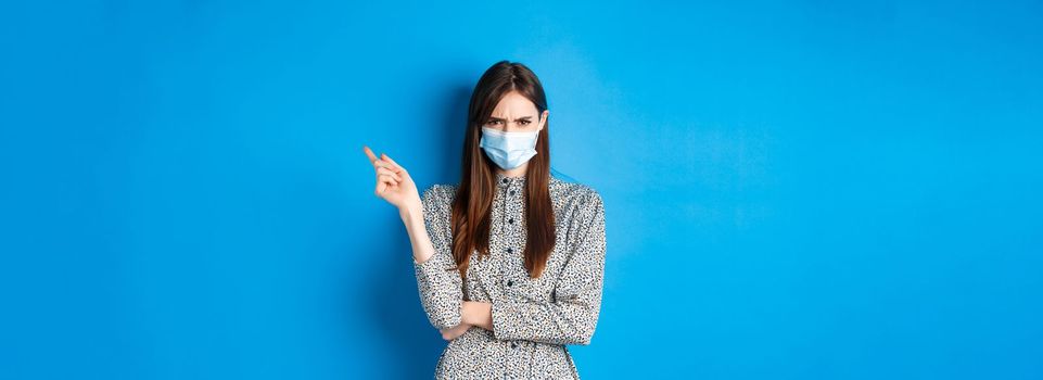 Covid-19, social distancing and healthcare concept. Angry young woman in medical mask frowning, pointing right at bad thing, condemn non-maskers, blue background.