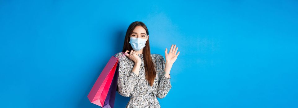 Covid-19, pandemic and lifestyle concept. Friendly cute woman wear medical mask in shopping mall, waving hand and holding bags, say hello, standing on blue background.