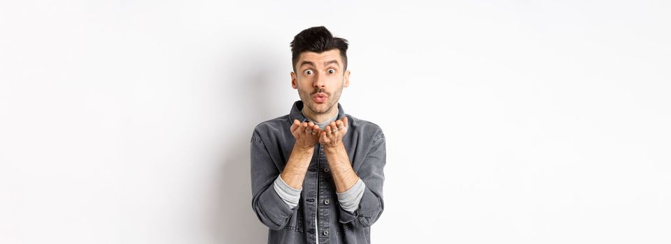 Romantic handsome man blowing air kiss, looking silly at camera, standing in trendy clothes on white background.