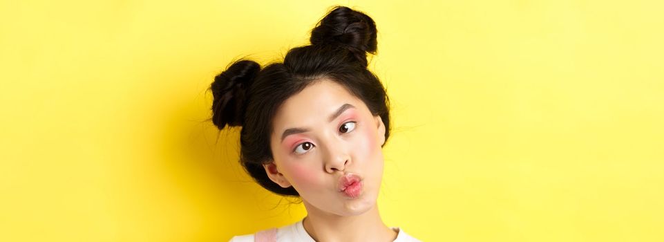 Close up portrait of funny asian teen girl with glamour makeup and hairstyle, squinting eyes and pucker lips silly, standing on yellow background.