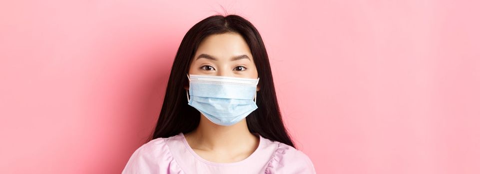 Covid-19 and healthy people concept. Close-up of asian cute girl wearing medical mask during virus outbreak, standing against pink background.