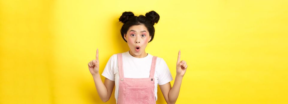 Excited asian female model with glamour makeup, pointing fingers up and say wow amazed, checking out promo deal, standing on yellow background.