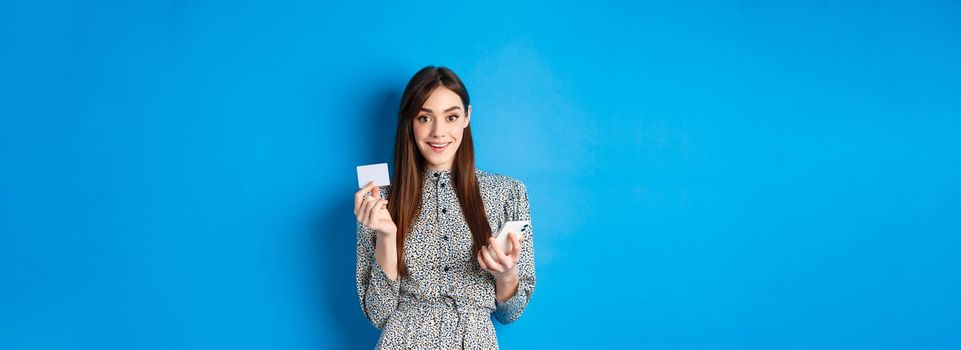 Online shopping. Beautiful woman paying for order with smartphone and plastic credit card, smiling at camera, wearing dress, blue background.