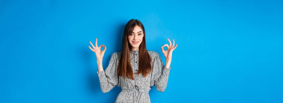 Confident adult woman in dress with natural hair color, showing okay signs and smiling, like and approve something good, standing on blue background.