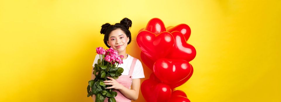 Happy Valentines day. Beautiful asian woman dressed for romantic date, holding bouquet of flowers and smiling. Girl with roses standing near heart balloons, yellow background.