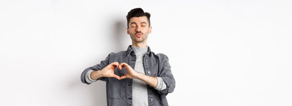 Cute young man waiting for kiss and showing heart gesture, confess on Valentines day, I love you sign, standing against white background.