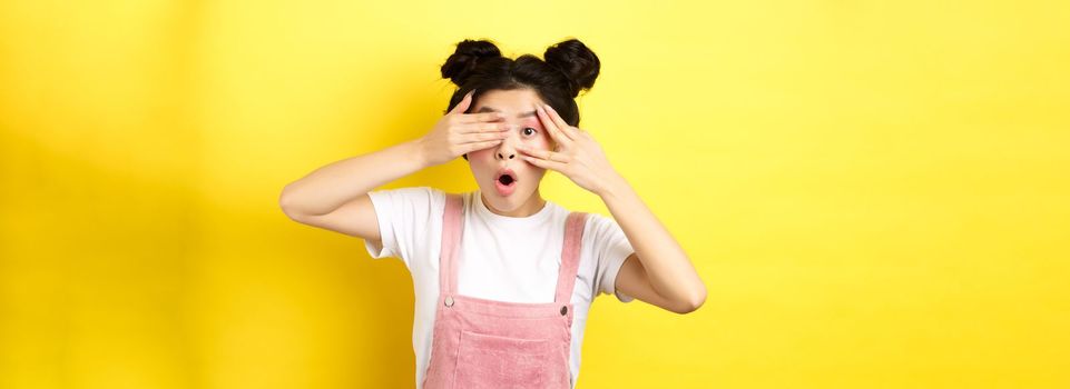 Excited asian teen girl covering eyes with hands and peeking through fingers, gasping amazed at camera, standing in summer clothes on yellow background.