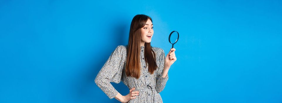 Excited girl looking left with magnifying glass, found interesting promo, investigating or searching, standing on blue background.