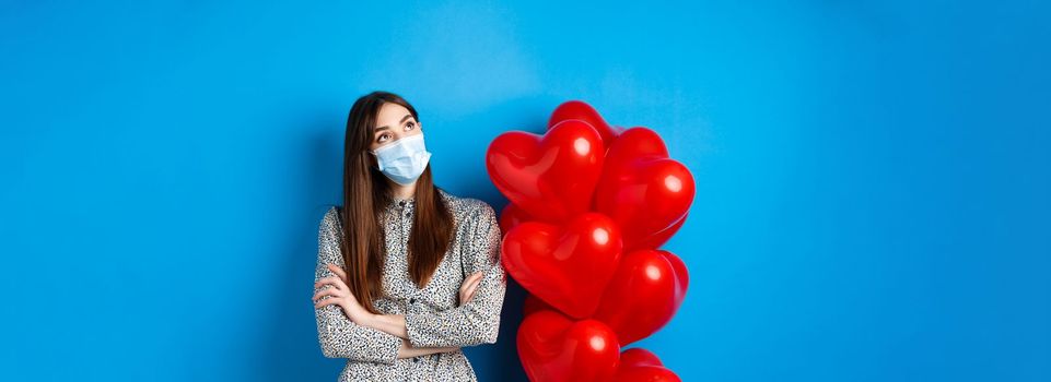 Covid-19, quarantine and health conept. Dreamy beautiful girl in face mask and dress, looking at upper left corner pensive, standing near Valentines day balloons, blue background.