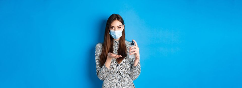 Covid-19, social distancing and healthcare concept. Beautiful girl in medical mask using hand saniziter, holding antiseptic bottle, standing on blue background.