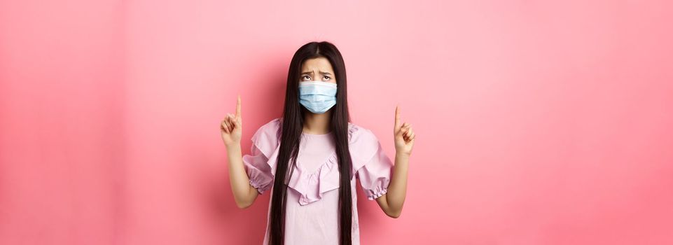 Coronavirus, quarantine and lifestyle concept. Disappointed sad asian girl pointing fingers up, frowning and sulking at unfair situation, standing against pink background.