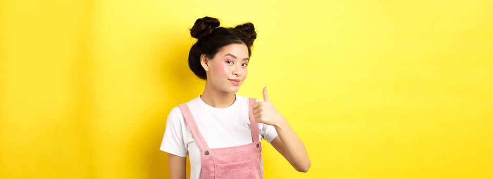 Beautiful asian girl with bright pink makeup, showing thumb up in approval and smiling satisfied, recommending promo, yellow background.