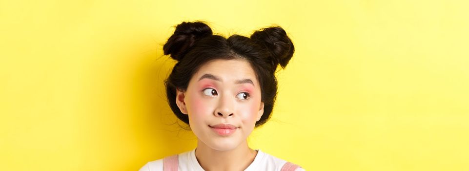 Close-up portrait of cute glamour asian girl, bright makeup and hairstyle, looking aside at logo with silly face, yellow background.