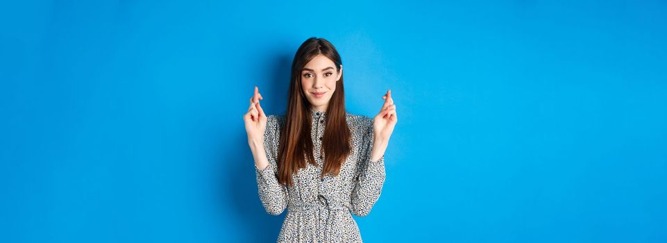 Hopeful smiling woman cross fingers for good luck and looking positive, praying or making wish, standing on blue background.