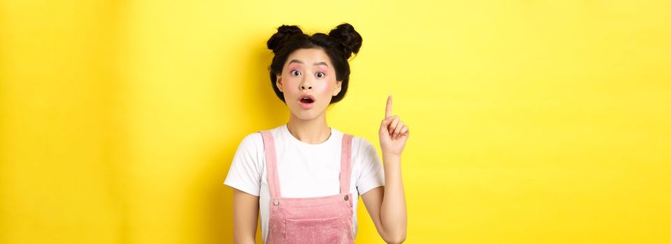 Excited teen asian girl having idea, raising finger and say suggestion, wearing stylish summer clothes with makeup, yellow background.