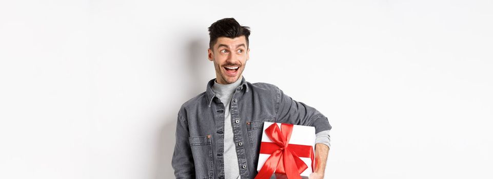 Cheerful handsome guy looking at empty space with amazed smile, checking out special valentines day offer, holding big romantic gift box, standing on white background.