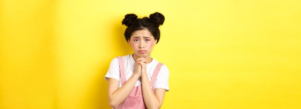 Sad asian girl pleading for help, begging you, sulking and whining, standing on yellow background.