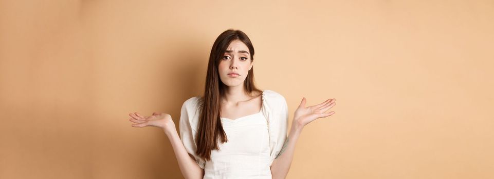 I dont know. Clueless sad girl shrugging shoulders and looking unaware, have nothing to say, standing on beige background.