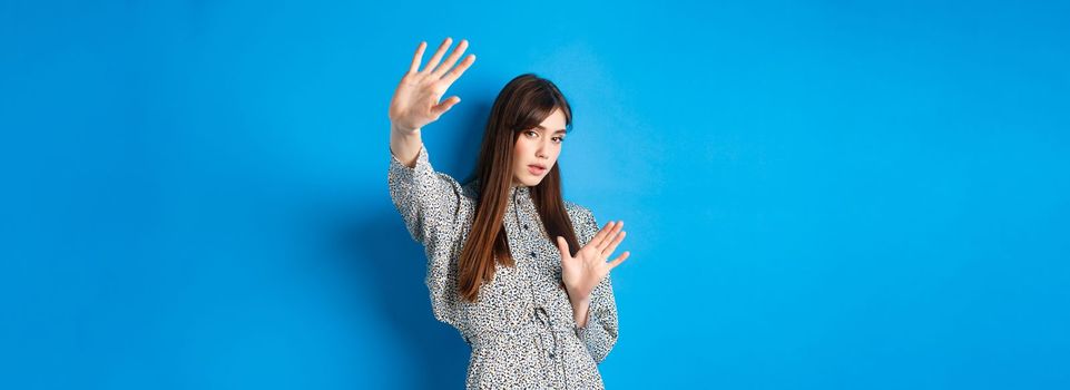 Image of young woman raising hands up defensive, covering herself from camera fleshlights, standing on blue background. Copy space