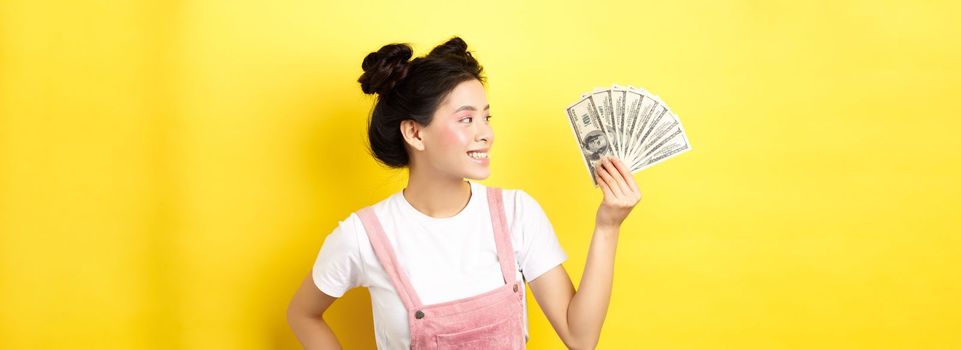 Shopping. Rich and stylish asian female model showing money, looking at dollar bills with pleased smile, standing on yellow background.