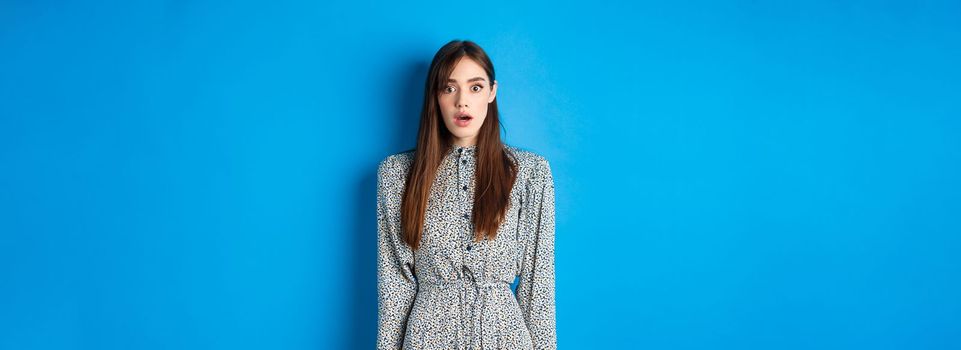 Shocked cute girl in dress looking at camera with opened mouth, gasping startled, see something shocking, standing on blue background.