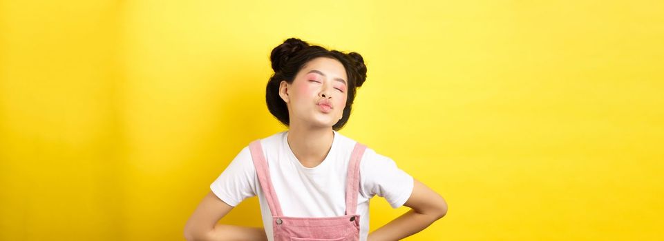 Romantic korean girl close eyes and pucker lips, waiting for sweet kiss on Valentines day, kissing lover, standing on yellow background.