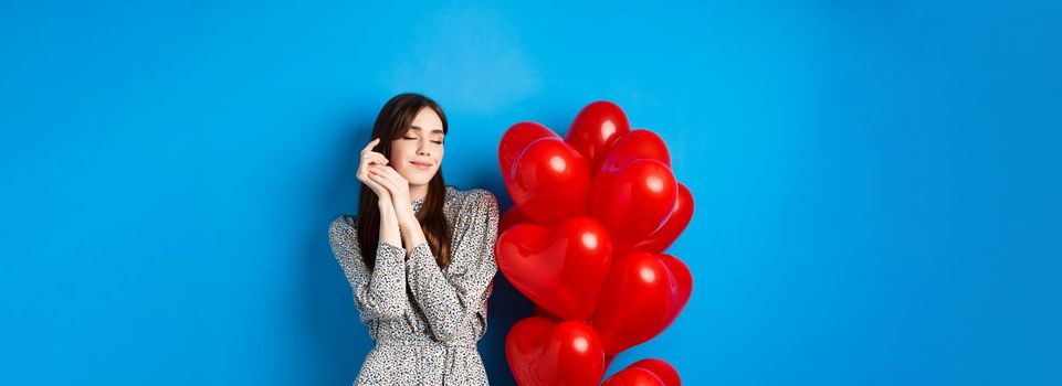 Valentines day. Dreamy romantic woman close eyes and imaging lovely date, standing near heart balloons and smiling, blue background.