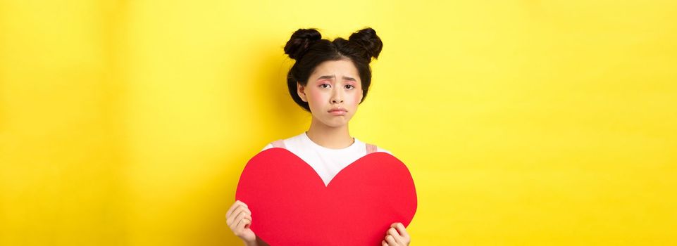 Heartbroken asian woman showing big red heart cutout and looking sad, feeling lonely on lovers day, showing valentine card at camera, yellow background.