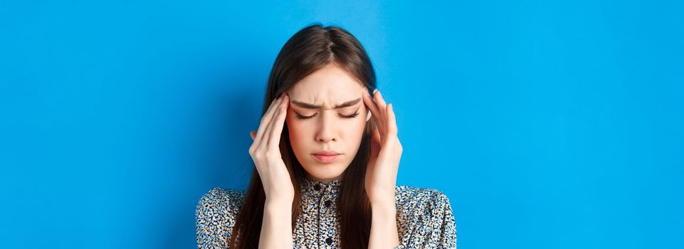 Close-up portrait of young woman feel sick, touching head temples and frowning from headache, having migraine, standing on blue background.