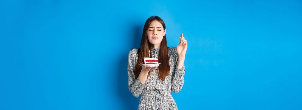 Hopeful birthday girl wish for dream come true, cross fingers with eyes closed, holding cake with candle, blue background.