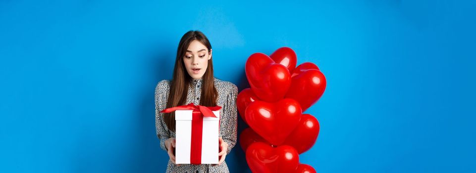 Valentines day. Image of beautiful girl looking surprised at gift box, receive present from lover, standing in dress on blue background.