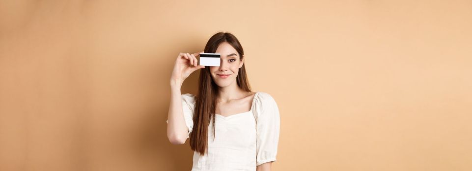 Smiling pretty girl showing plastic credit card over eye and looking satisfied, standing on beige background. Concept of shopping.
