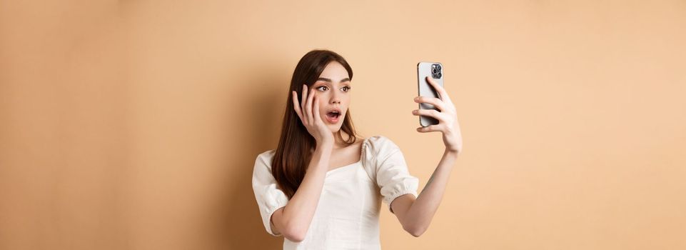 Fashion girl record smartphone blog, taking selfie on cellphone, standing on beige background.