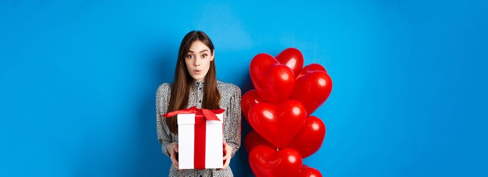 Valentines day. Surprised attractive girl looking amazed at camera, holding big romantic gift, standing near red hearts balloons, blue background.