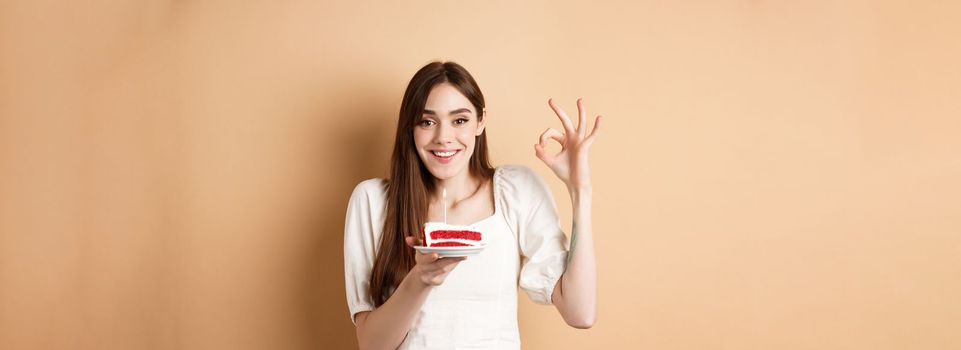 Happy birthday girl show okay gesture and hold bday cake, making wish on her holiday, standing on beige background.