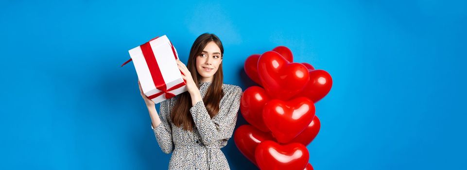Valentines day. Beautiful woman shaking gift box to guess what inside, celebrating lovers holiday, standing near red hearts, blue background.