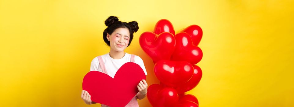 Happy Valentines day. Silly and beautiful asian woman smiling dreamy, showing red heart, imaging romantic date with lover, standing on yellow background.