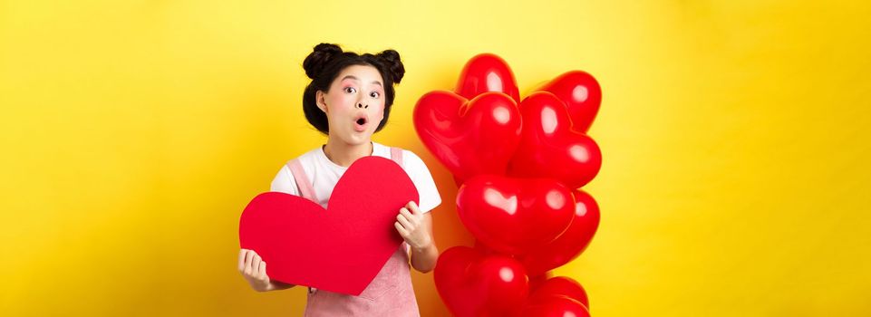 Happy Valentines day. Silly asian girl showing big red heart postcard and looking amazed, standing near red romantic balloons, yellow background.