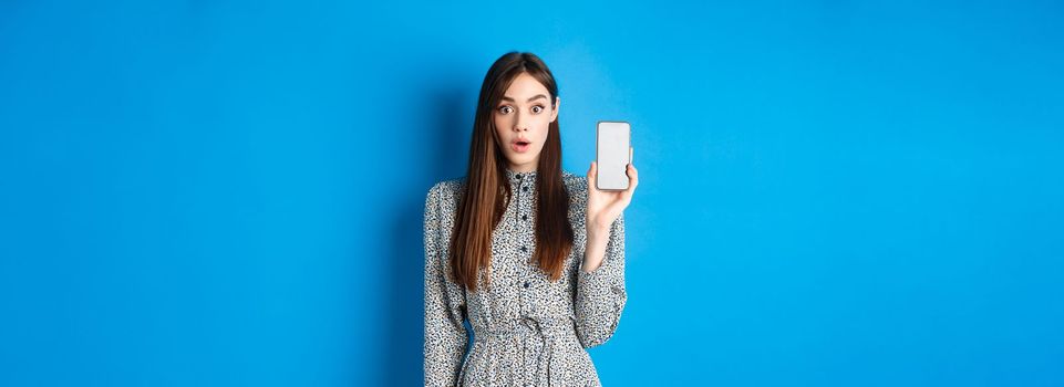 Excited young woman in dress, gasping amazed, showing empty smartphone screen and look impressed at camera, blue background.