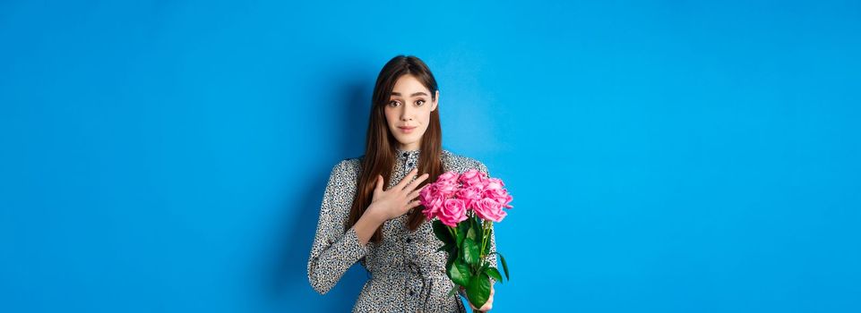 Valentines day concept. Touched and thankful pretty girl, holding hand on heart and smiling grateful for gift, holding bouquet of roses, standing on blue background.