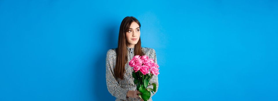 Valentines day concept. Pretty romantic girl looking dreamy at empty space, holding bouquet of pink roses on date, standing alone on blue background.