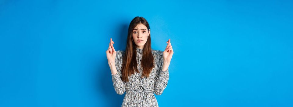 Nervous young woman in dress cross fingers and biting lip worried, praying or making wish, standing on blue background.